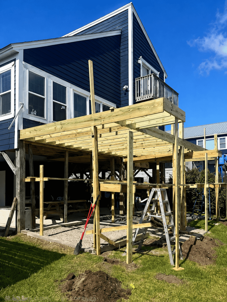 Editorial illustration of elevated deck framing beside blue horizontal siding and white-trimmed windows.